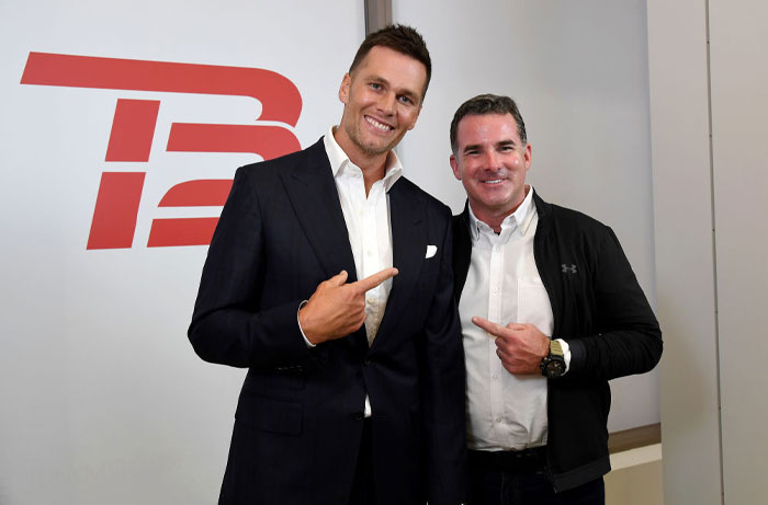 Tom Brady, dressed in a suit, smiling and pointing to a man beside him in casual attire, both posing in front of a "TB12" logo backdrop.