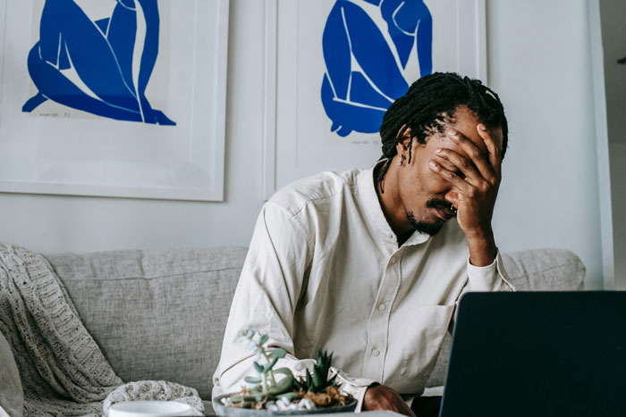 Man looking stressed in front of a laptop, sitting on a couch, with hand on face.