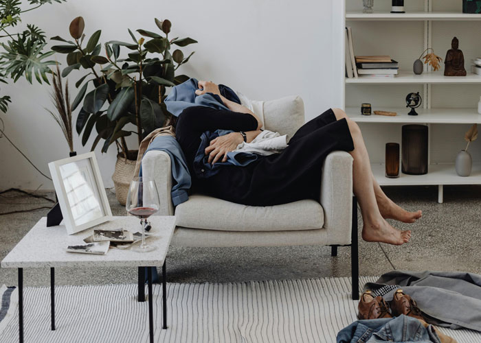 Couple relaxing on sofa, surrounded by plants and decor, with wine glass and scattered clothes, reflecting on their relationship.