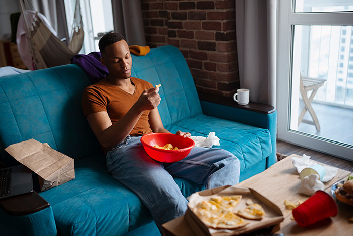 Man in an orange shirt sits on a blue couch, eating from a red bowl, highlighting roommate dynamic issues.