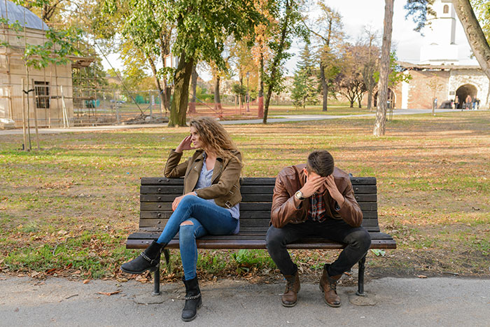 Woman upset on park bench, man covering face, tension over friend's housewarming party invitation.