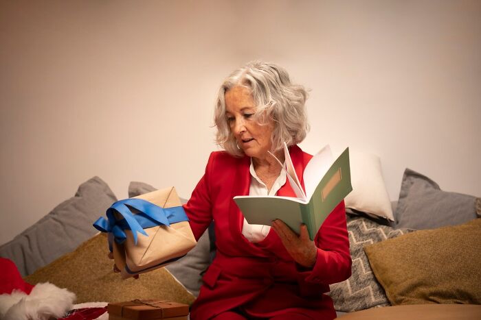 Woman in red suit checking a gift with a blue ribbon while holding a green notebook.