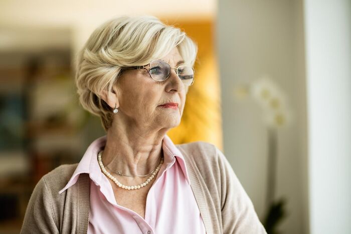 Elderly woman with glasses and pearl necklace looking contemplative.