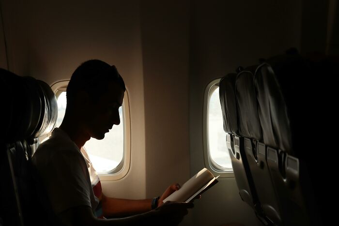 Passenger reading on a long flight, experiencing discomfort in an airplane seat, with dim lighting.