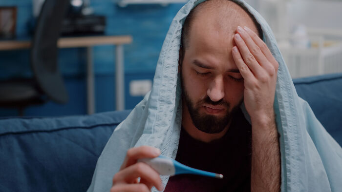 Man feeling unwell, wrapped in a blanket, checking a thermometer after worst experience on a long flight.