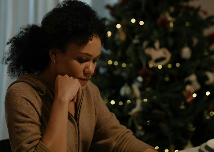 Woman looking thoughtful near a Christmas tree, capturing holiday stress as one of the worst things about Christmas.