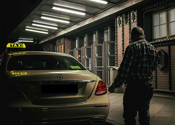 Man standing near a taxi at night, under bright lights, in an urban setting.