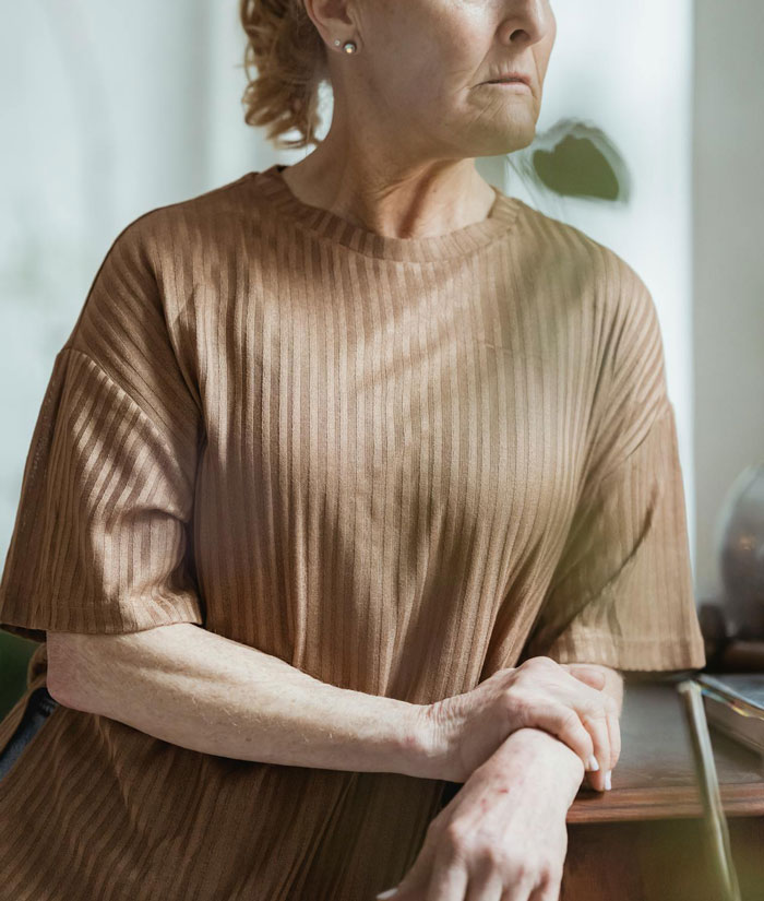 Elderly woman in a brown shirt, looking to the side thoughtfully, with hands resting on a table, related to pretend break-up.