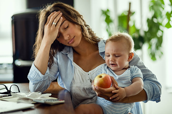 Stressed woman with baby, pressured by family for childcare at wedding, looks overwhelmed.