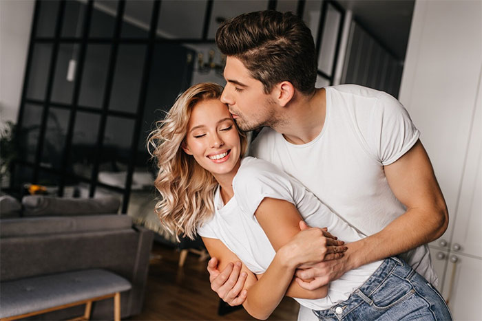 Woman in a happy embrace with boyfriend at home, wearing casual white shirts, smiling and surprised.