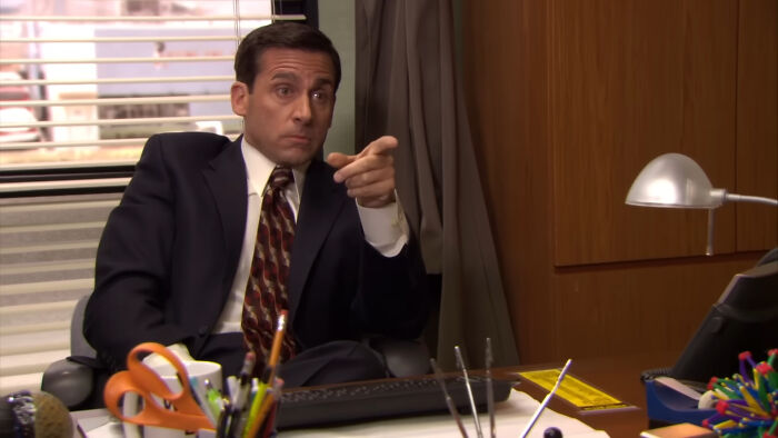 A man in a suit points while sitting at a desk cluttered with office supplies, a scene from a beloved comedy show.