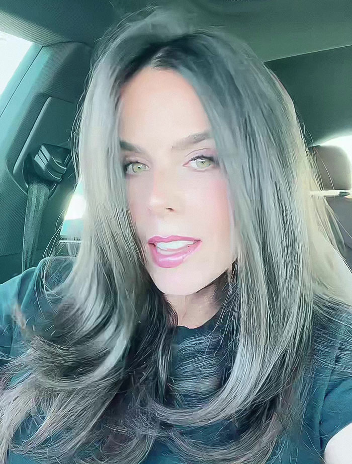 A woman with long hair, sitting in a car, looking directly at the camera.