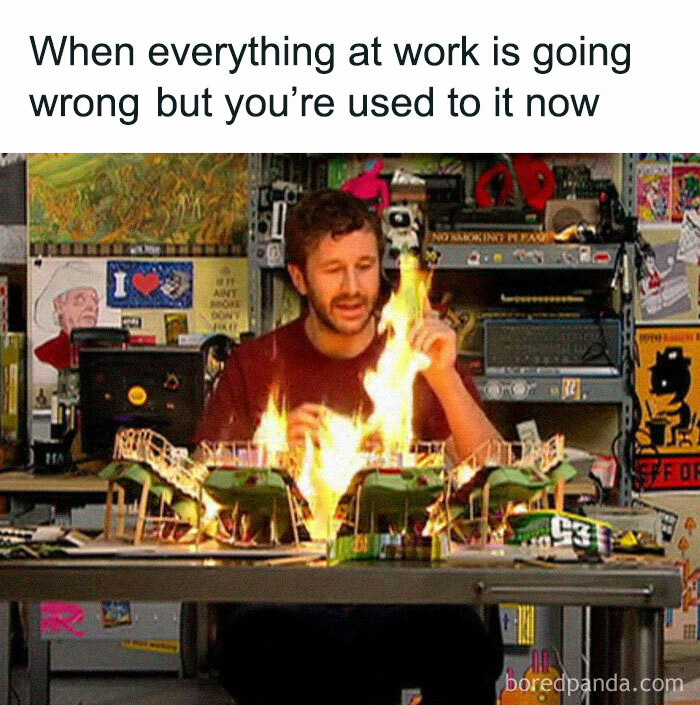 Man in office humorously reacting to a desk fire, surrounded by posters and office items, illustrating funny office memes.