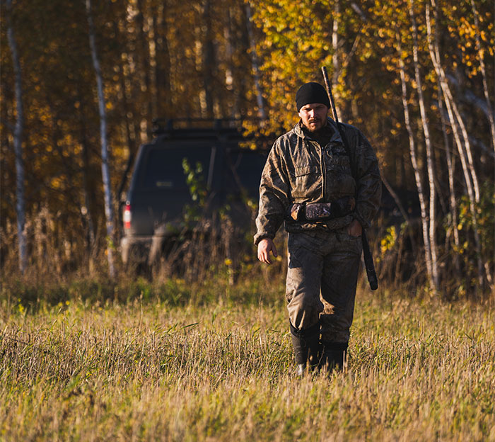 Man in camouflage gear walks through a field with a rifle, near a vehicle, during a deer hunt on private land.