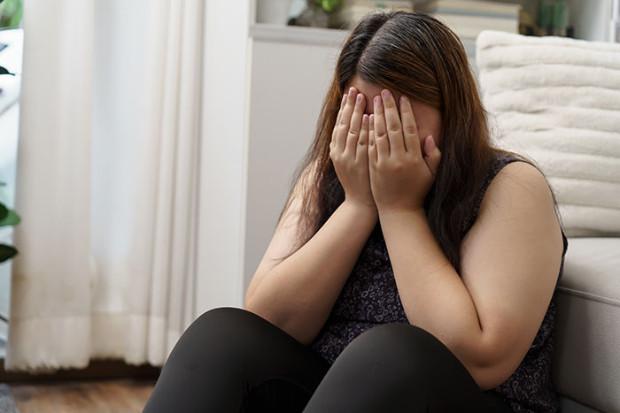 A woman sitting on the floor with her face in her hands, expressing emotions related to body image and being plus-size.