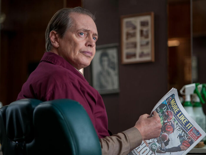 Actor holding a newspaper, sitting in a chair, looking contemplative.