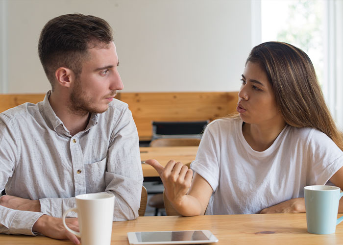 A Canadian woman in a white shirt gestures while talking to a man in a bright room.