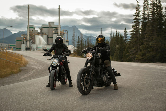 Two motorcyclists in dark gear on a road near an industrial area, under a cloudy sky, depicting themes of corruption.