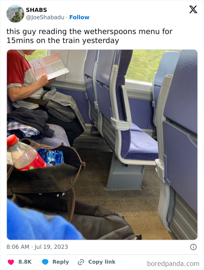 Man humorously reading a menu on a train, surrounded by seats and a bag with a drink inside.