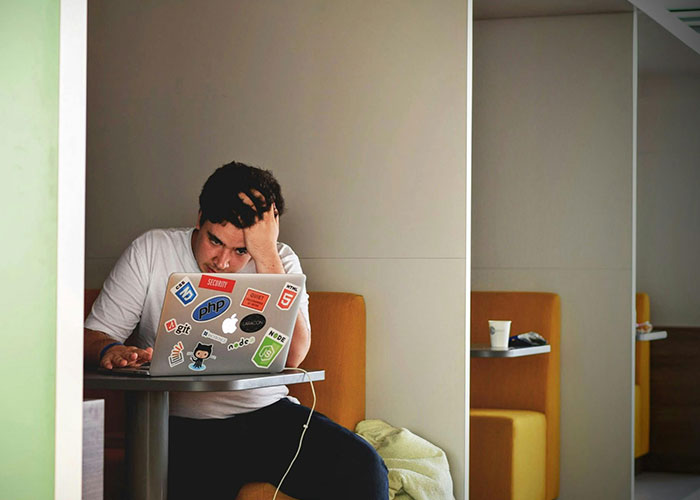 Person looking stressed with hand on forehead, sitting in cubicle, using a sticker-covered laptop; awkward job interview vibe.