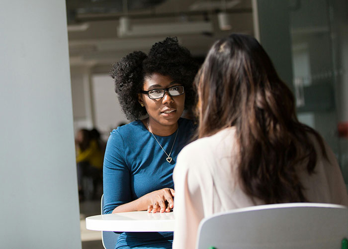 Two women in a job interview setting, one wearing glasses and blue dress, talking over a round table.