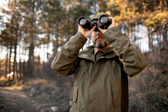 Man in green jacket looking through binoculars in forest, hunting deer on private land.