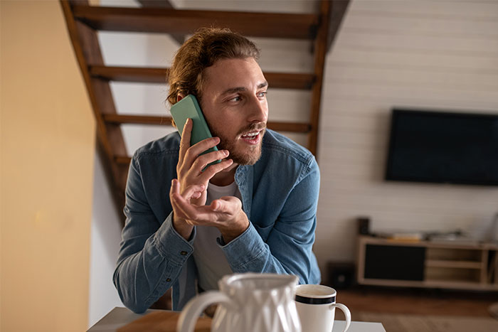 Man in blue shirt talking on phone, discussing last-minute party cancellation at home.