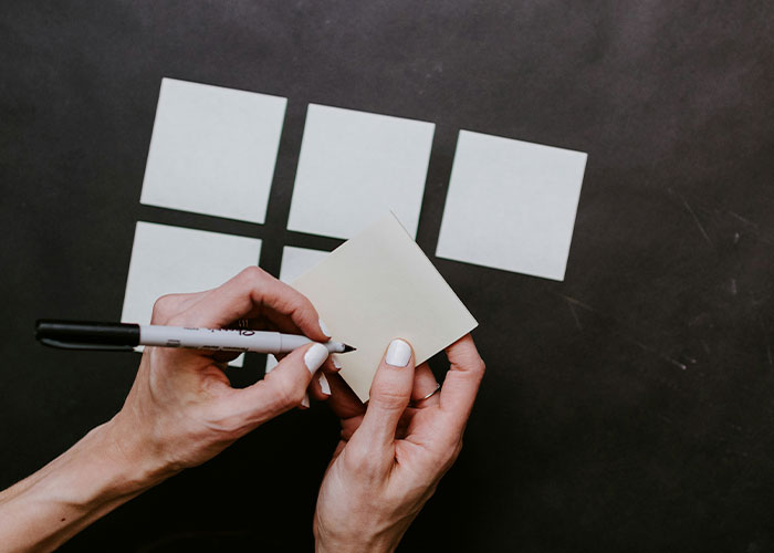 A person writes on a sticky note with a marker, surrounded by more sticky notes on a dark surface.