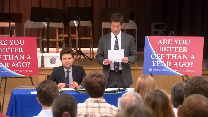 Two men in suits at a public meeting with signs saying, "Are you better off than a year ago?"