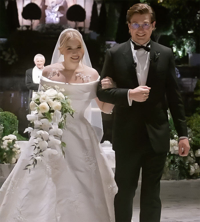 Bride in viral wedding, smiling in white gown, holding groom's arm, surrounded by floral decor and officiant in background.