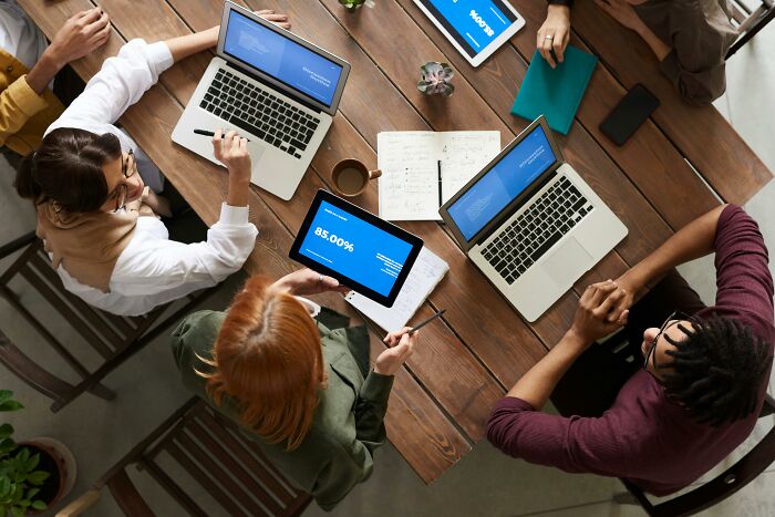 A group discusses unethical life hacks over laptops and tablets at a wooden table.