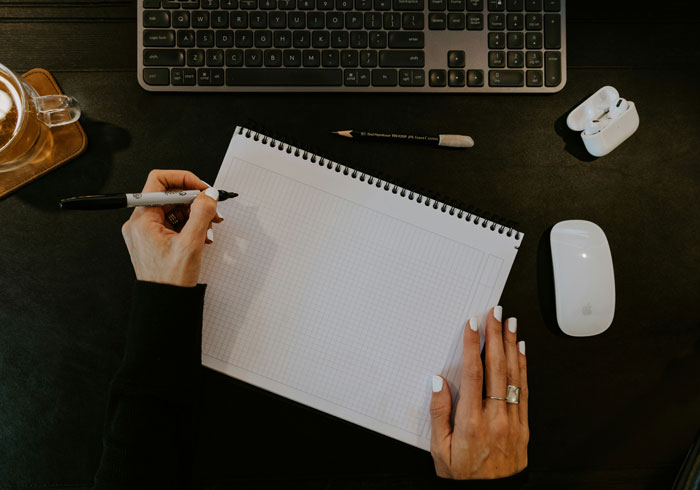 Person writing on notepad at desk with keyboard, using quality hacks for real-life.