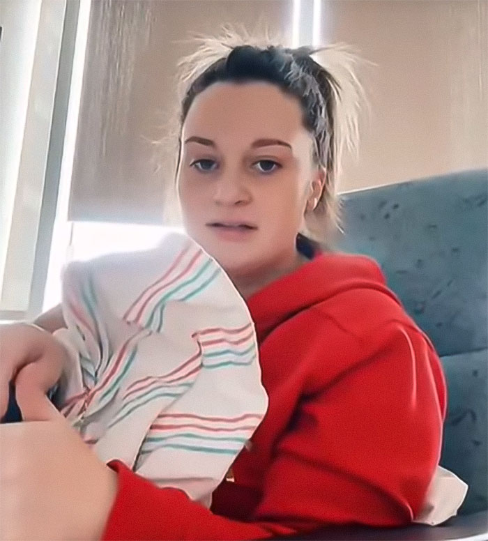 Woman in a red hoodie holding a baby wrapped in a striped blanket, sitting indoors.