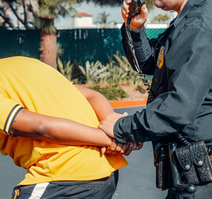 Officer handcuffing a person wearing a yellow shirt outdoors, holding a radio.