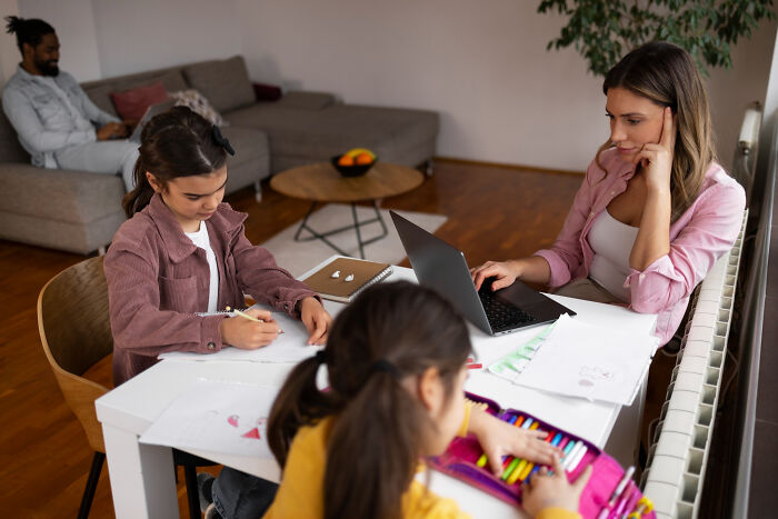 A parent working on a laptop at a table with children drawing, highlighting signs of a horrible parent.