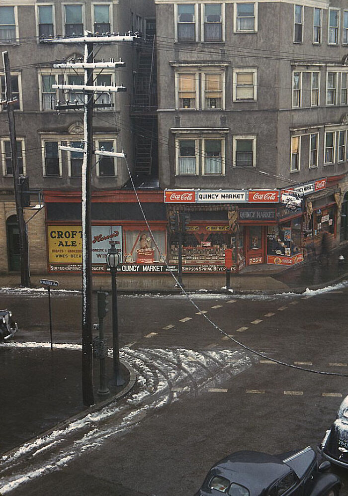 1940 color photo of a snowy urban street corner with a market, vintage cars, and buildings in the background.