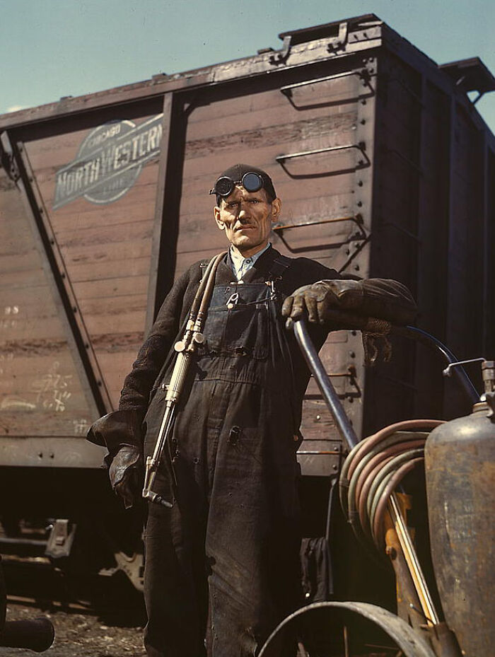 Worker in goggles and overalls stands by train car from 1940s, captured in vivid color photography.