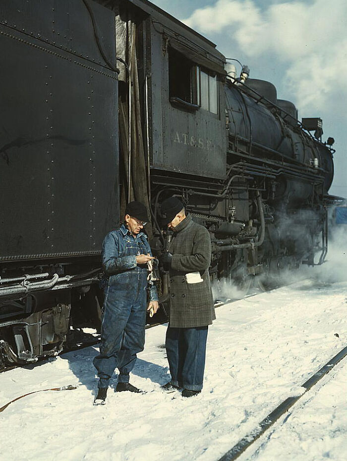 Two men in 1940s attire standing by a steam train on snow-covered tracks, captured in color photos.