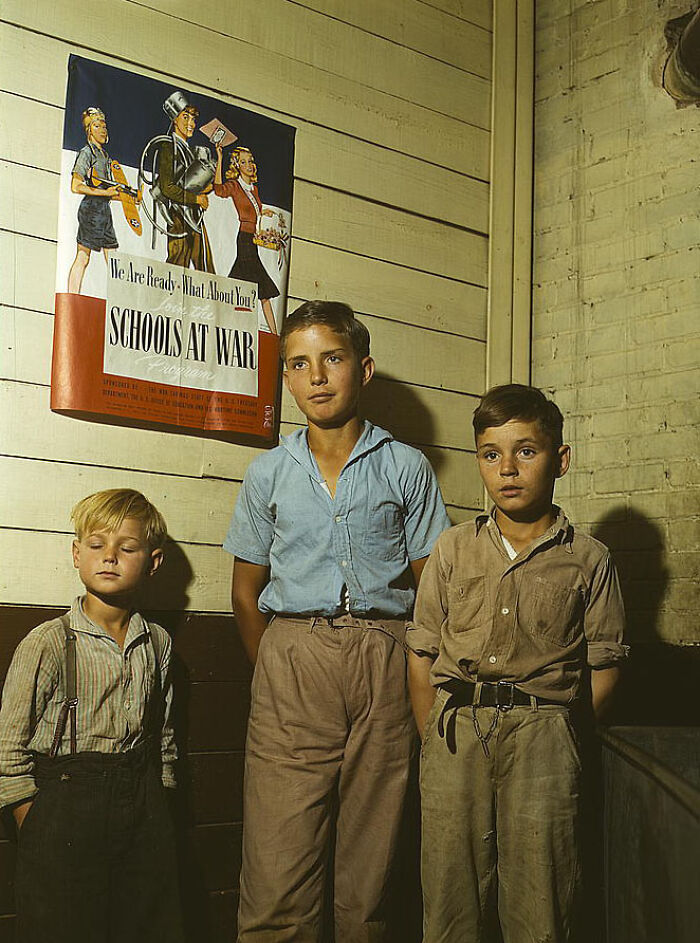 Three boys in front of a "Schools at War" poster, 1940s color photo showcasing wartime efforts.