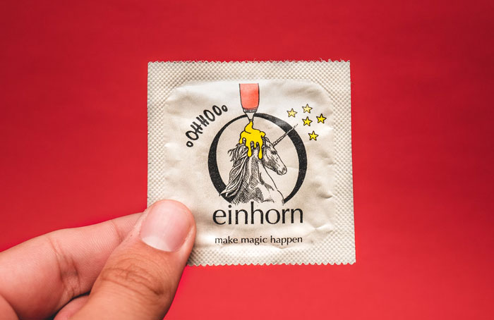 Hand holding einhorn condom with unicorn design, showcasing a quirky life hack product against a red background.