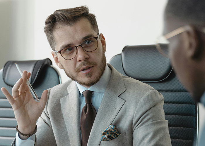 Man in a formal suit discussing awkward job interviews, holding a pen, in a boardroom setting.