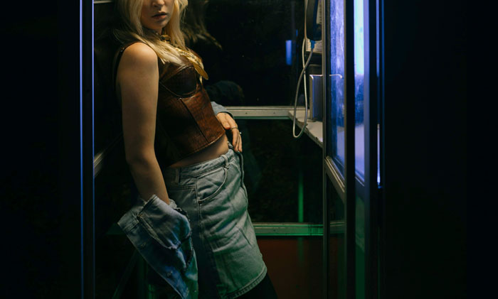 Person in dimly lit phone booth, wearing a denim skirt and jacket, portraying a mysterious, introspective scene.