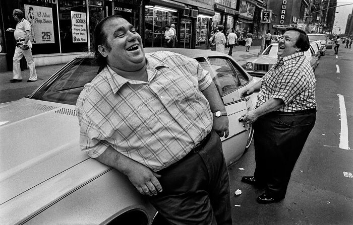 Two people laughing and leaning on a car in a busy city street, capturing an interesting moment.