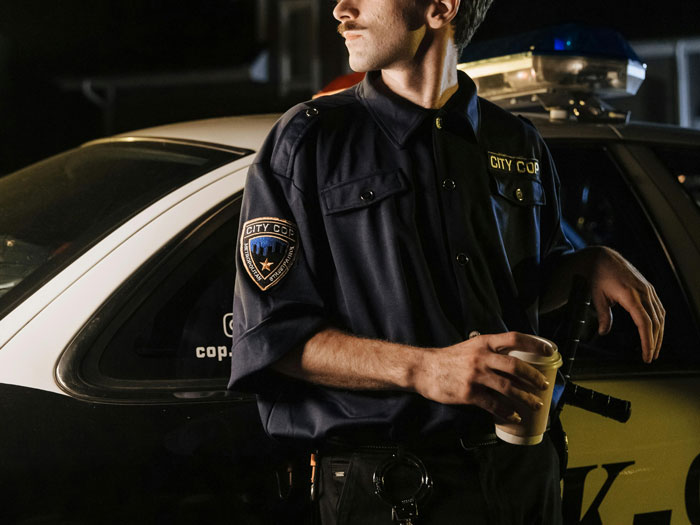 Officer in uniform leaning on police car, holding coffee.