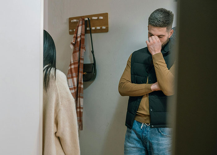 A tense couple stands in a hallway, the man looking frustrated and the woman facing away.