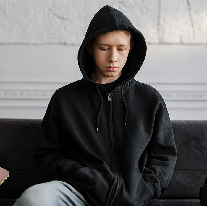 Teen in a black hoodie sitting on a sofa, looking down and appearing contemplative.