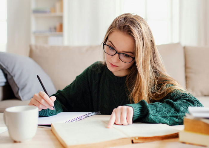 Teen girl with glasses studying, wearing a green sweater, writing in a book.