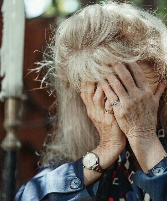 Elderly woman covering her face with hands, appearing distressed in a garden setting.