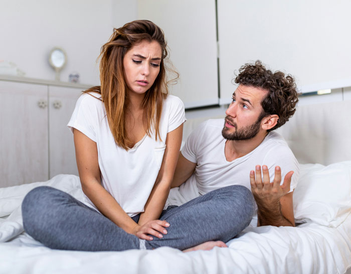 Confused couple sitting on a hotel bed, discussing vacation plans, expressing concerns about a MIL tagging along.