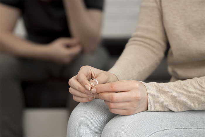 A woman holding a ring, sitting pensively on a couch with a second person in the background.